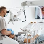 young-man-with-patient-bib-dental-chair-dentist-who-sits-him-he-looks-his-teeth-using-dental-microscope-holds-dental-bur-mirror-min